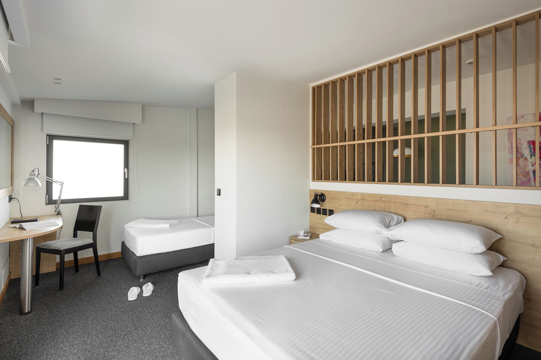 A hotel room with a large bed, a desk with a chair, a small bedside table, and a window.