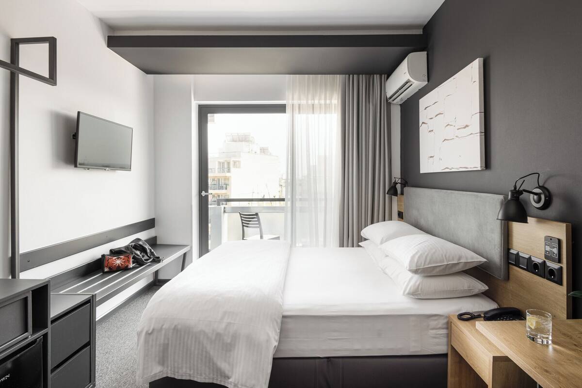A modern hotel room with a bed, bedside table, TV, and a view of the city through the window.