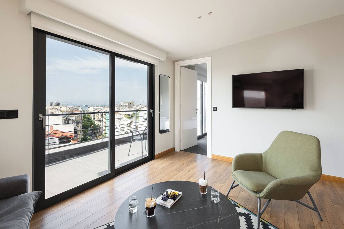 A modern living room with a large sliding glass door leading to a balcony, a flat-screen TV mounted on the wall, and a round coffee table with drinks and a book.