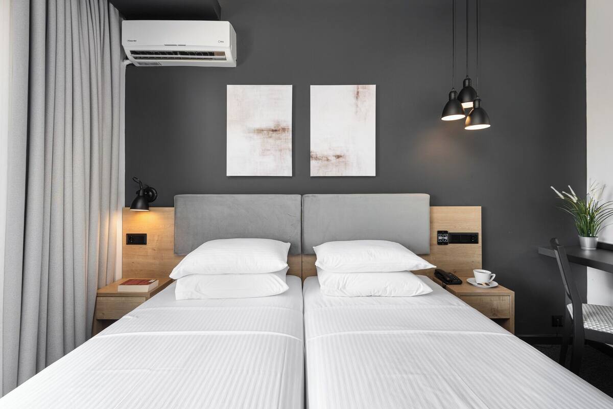 A modern hotel room with a grey headboard, two beds, a bedside table with a lamp, a phone, and a cup. There are two abstract paintings on the wall and an air conditioning unit above the bed.