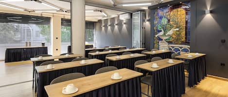 Un salón de conferencias con varias mesas, cada una con una taza y platillo blancos, y un ventanal con persianas.