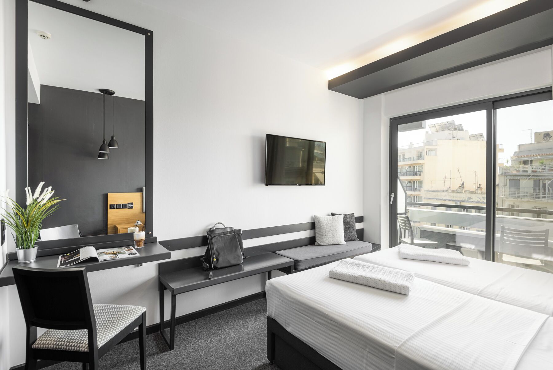 Een moderne hotelkamer met een groot bed, een bureau met stoel, een aan de muur bevestigde televisie en uitzicht op de stad door grote ramen.