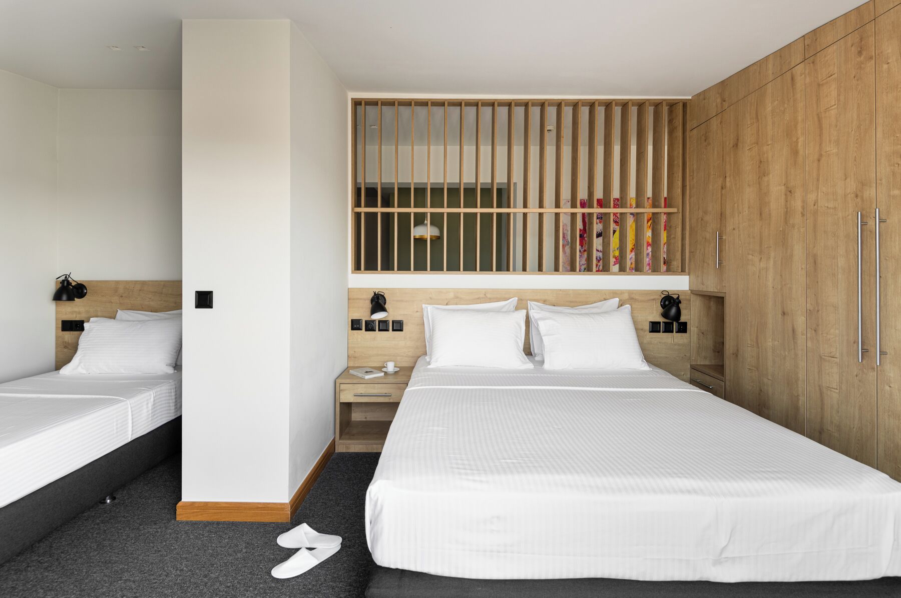 Habitación de hotel con dos camas individuales, un armario de madera, una mesita de noche con lámpara y una ventana con persianas verticales.