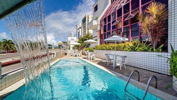 A swimming pool with a water feature, surrounded by outdoor seating and plants. - Happy Ponta Negra Express (Natal)