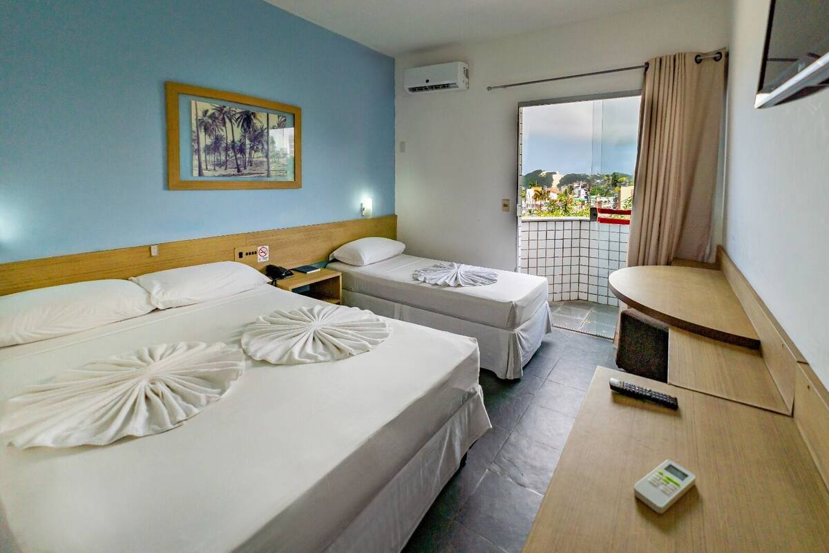 A hotel room with two beds, a small table, a chair, a window with a view of buildings, and an air conditioning unit.