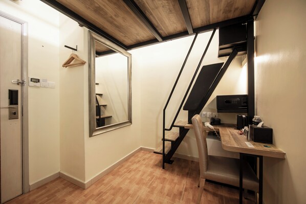 A modern room with a wooden floor, a desk with a chair, a hanging coat rack, a large mirror, and a staircase with a black railing.