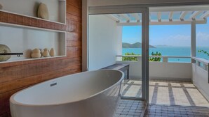 Een moderne badkamer met een vrijstaand bad, houten planken met decoratieve objecten, en een balkon met uitzicht op zee.