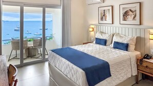 Hotelkamer met een groot bed, een balkon met uitzicht op zee, rieten meubilair en twee afbeeldingen in lijsten aan de muur.