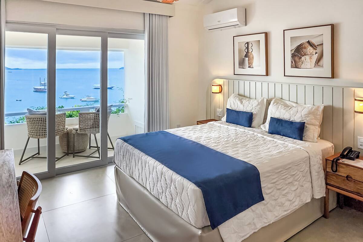 A hotel room with a large bed, a balcony with a view of the sea, wicker furniture, and two framed pictures on the wall.