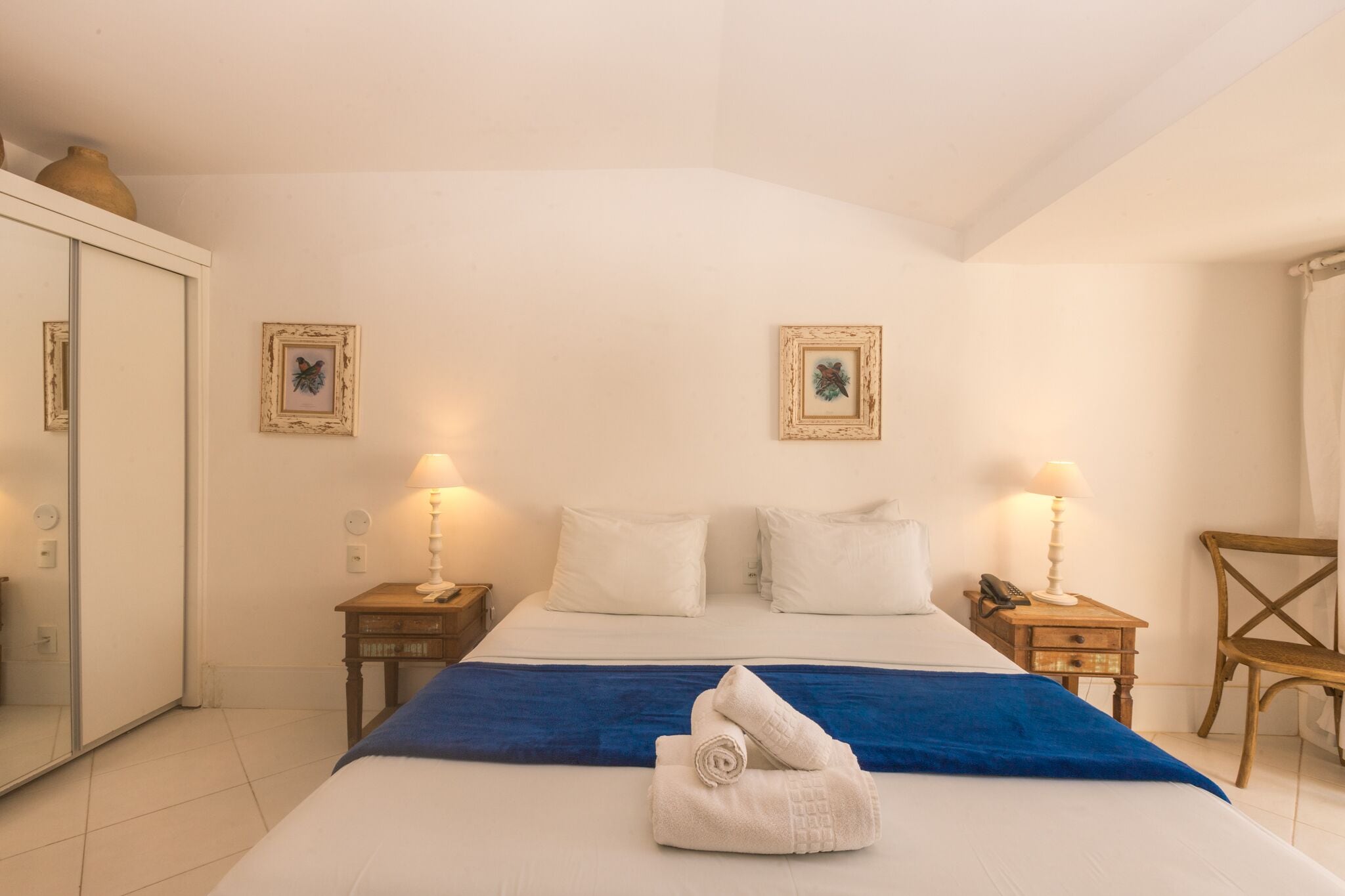 A hotel room with a bed, two bedside tables with lamps, a wooden chair, a wardrobe, and two framed pictures on the wall.