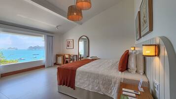 A hotel room with a large window offering a view of the sea and cruise ships, a bed with a red throw blanket, a bedside table with a remote control, and a wall-mounted light fixture.