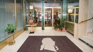 L’entrée d’un hôtel, avec un tapis où est écrit « BIENVENUE », un comptoir d’accueil et une vue sur le hall.