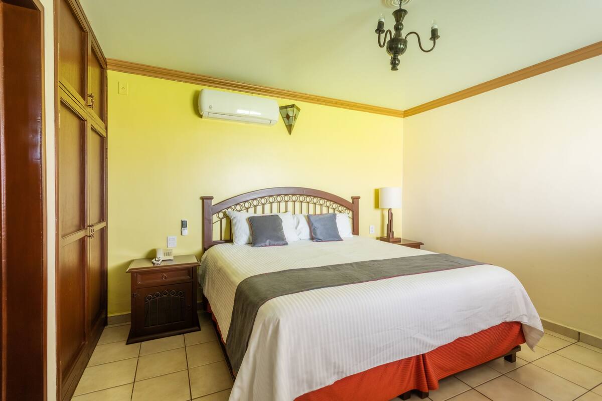 A hotel room with a large bed, wooden headboard, bedside tables, an air conditioning unit, and a chandelier.