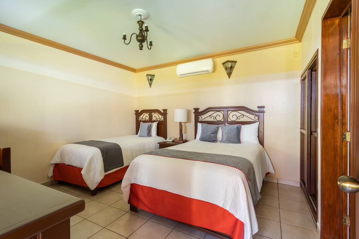A hotel room with two beds, a wooden headboard, a nightstand, a lamp, and a ceiling light fixture.