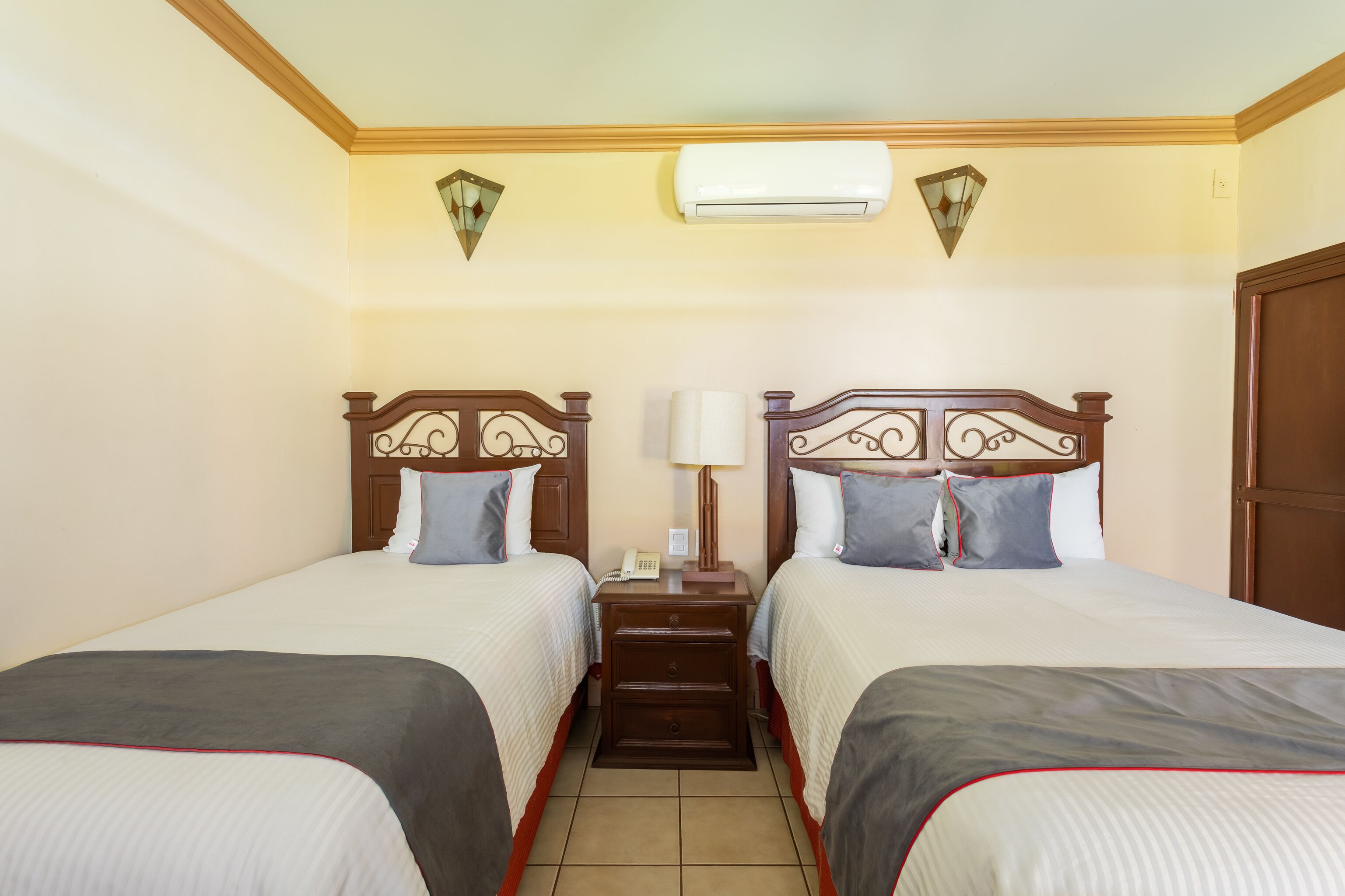 Een hotelkamer met twee enkele bedden, een nachtkastje met een lamp en een airconditioning.
