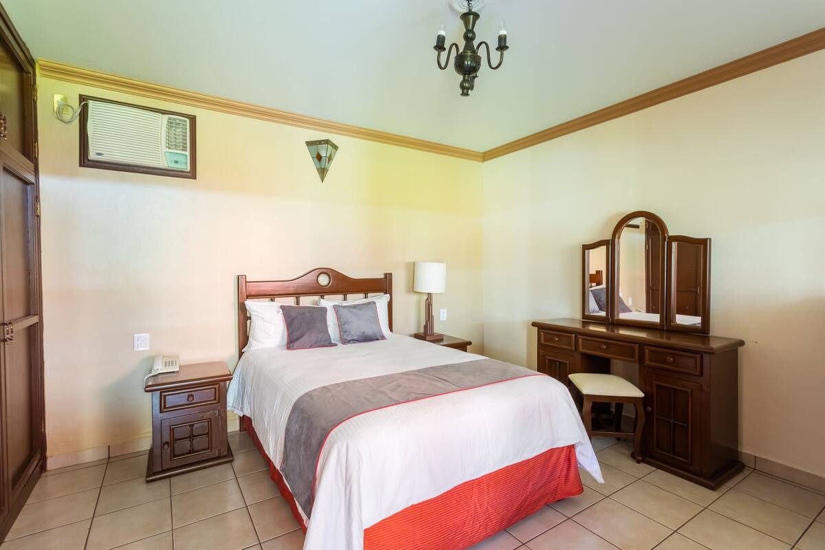 A hotel room with a bed, bedside table, lamp, dresser, mirror, and a wall-mounted air conditioning unit.