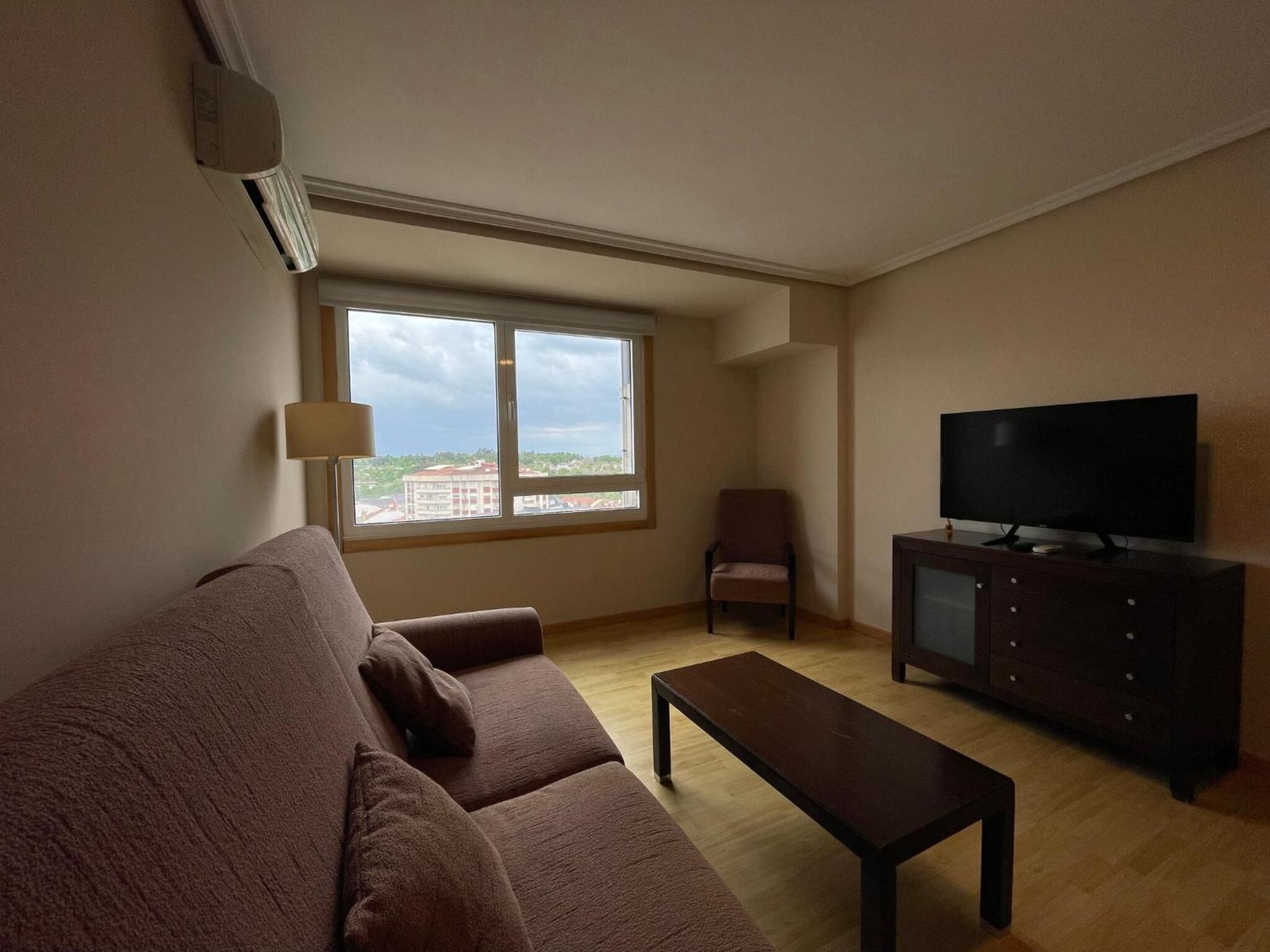 Una sala de estar con un sofá, un televisor sobre un mueble, una silla y una ventana con vistas a edificios.