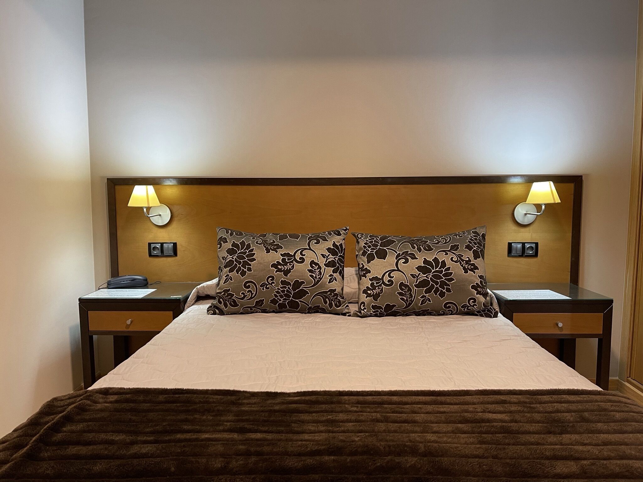 Une chambre d’hôtel avec un grand lit, deux lampes de chevet, une tête de lit en bois et un plaid de couleur marron foncé.