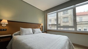 Une chambre d’hôtel avec un grand lit, deux lampes de chevet, une fenêtre donnant sur des immeubles et une tête de lit en bois.