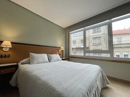 Habitación de hotel con una cama grande, dos lámparas de noche, una ventana con vistas a edificios y un cabecero de madera.