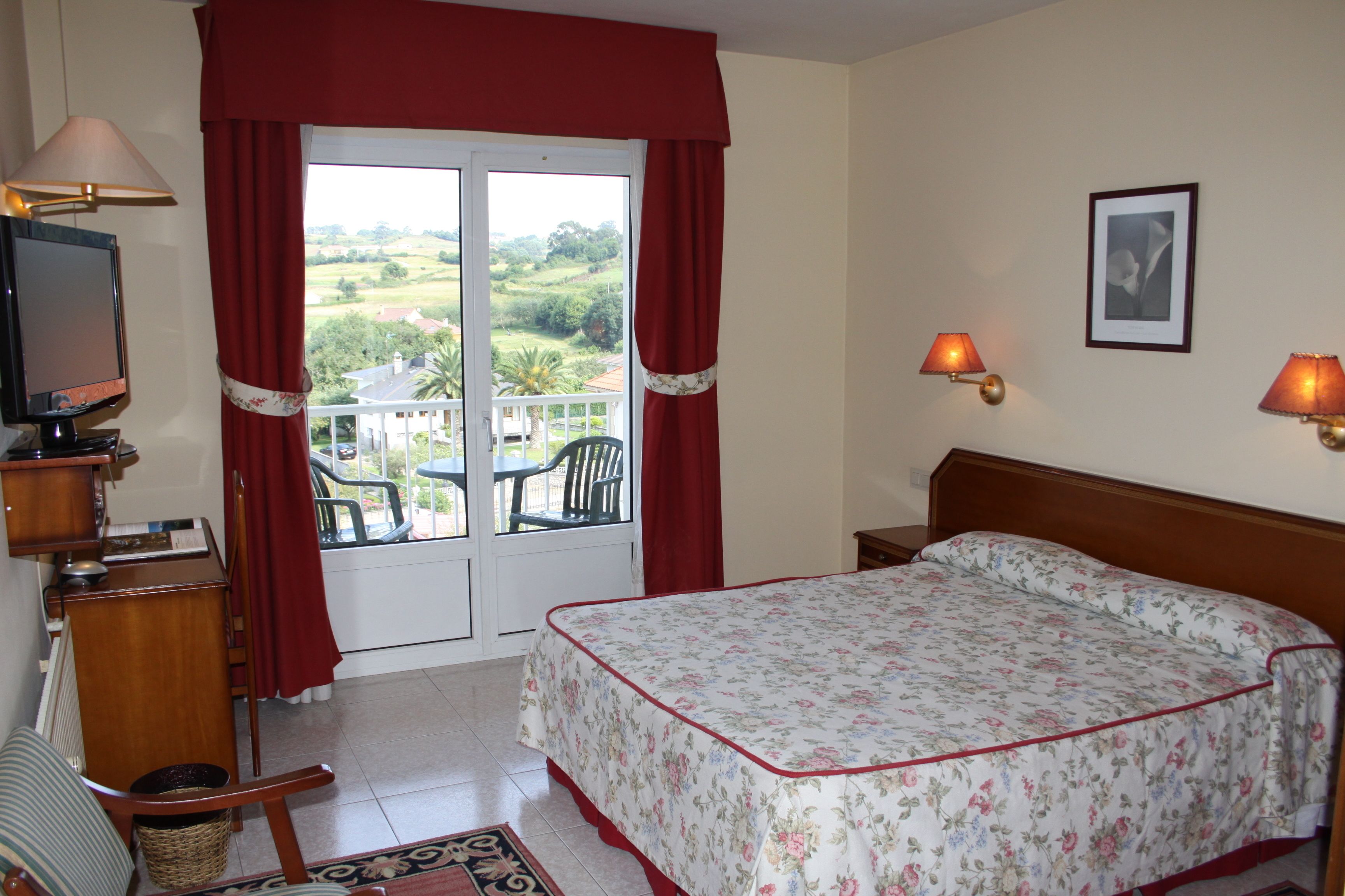 Una habitación de hotel con cama, televisión, escritorio, silla, ventana con cortinas y balcón con mesa y sillas.