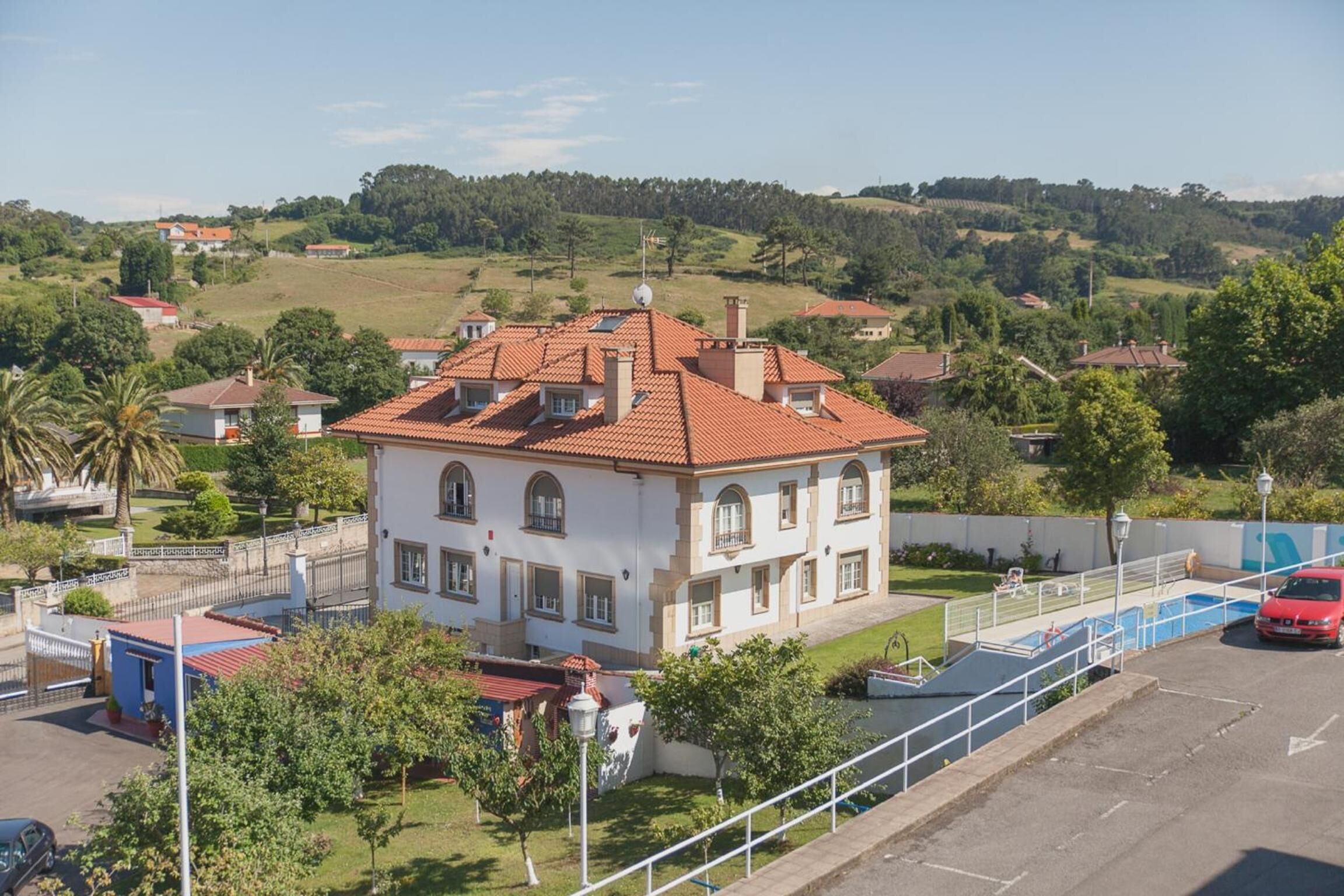 A two-story building with a red-tiled roof, surrounded by greenery and a swimming pool.