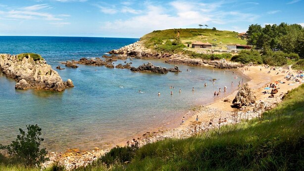 A coastal beach with rocky formations, clear blue water, and a grassy hill with buildings in the background. - El Carmen (Carreno)