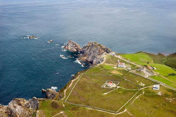 Vista aérea de una zona costera con acantilados rocosos, un faro y un pequeño conjunto de edificios con senderos y campos verdes.