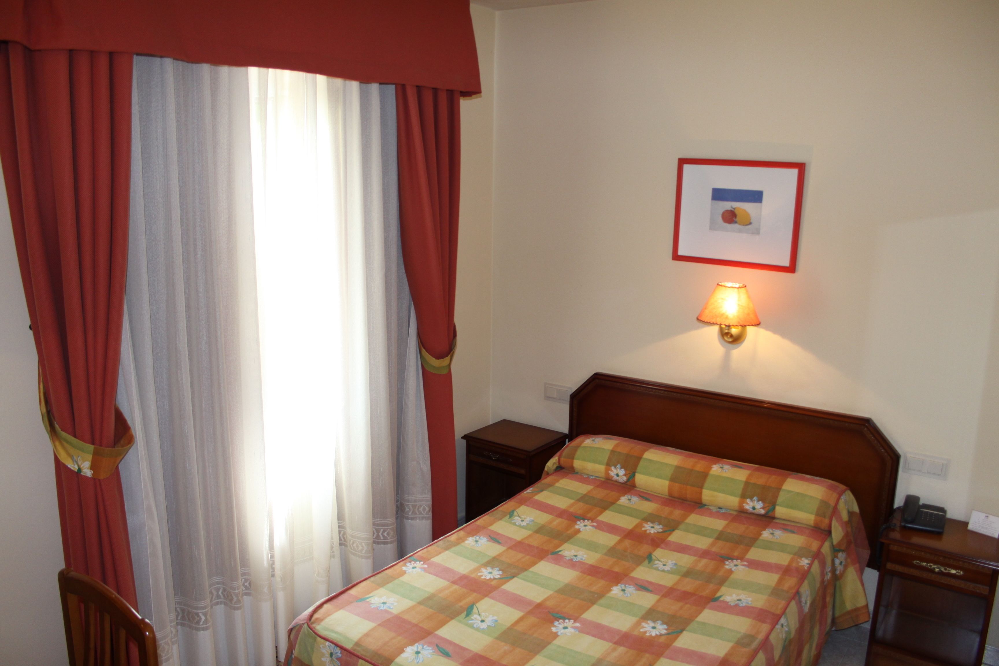 Ein Hotelzimmer mit Bett, Nachttisch, Lampe, Stuhl und einem Fenster mit Vorhängen.