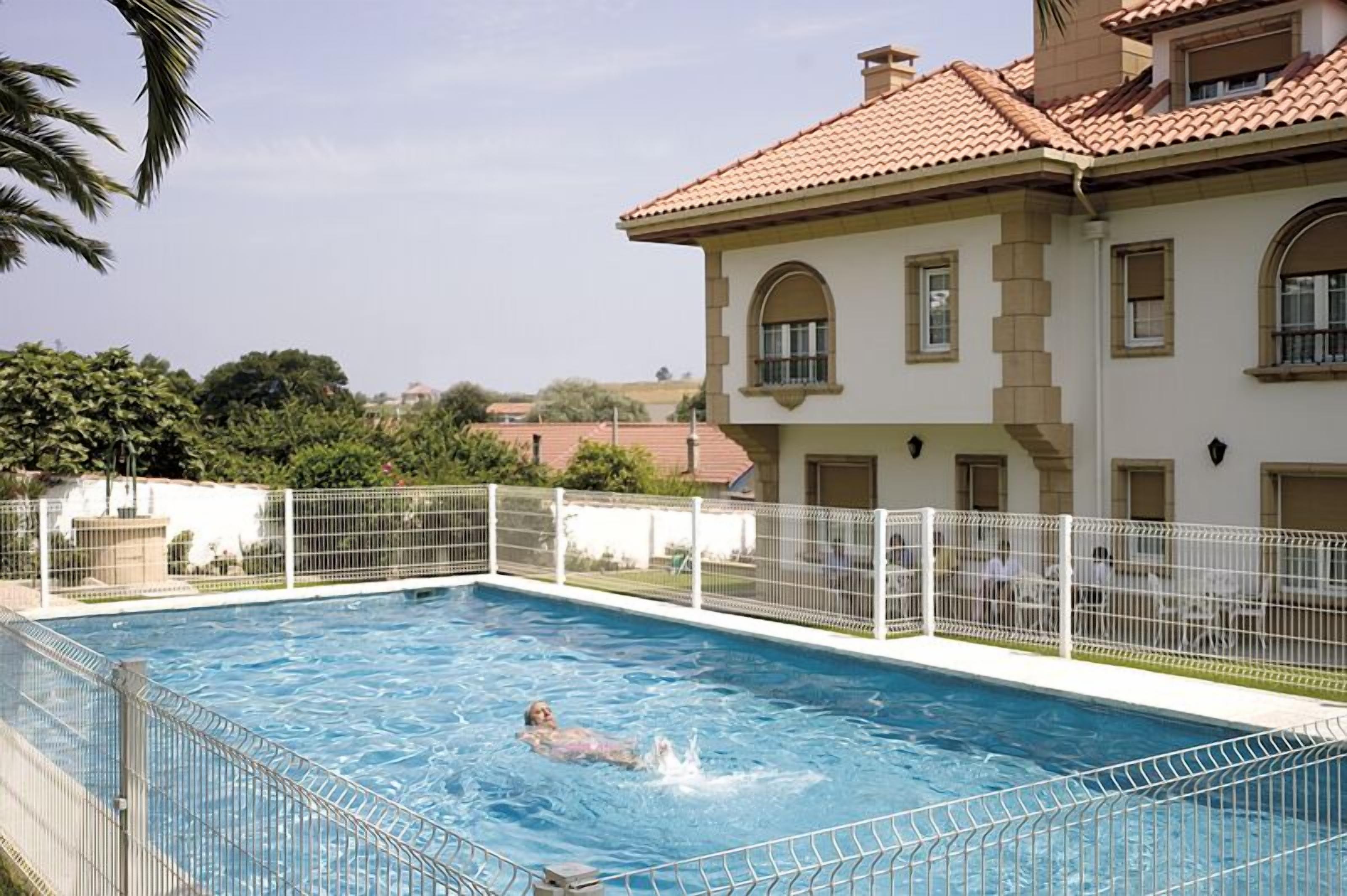 A swimming pool with a person swimming, surrounded by a white fence, and a building with a tiled roof in the background.