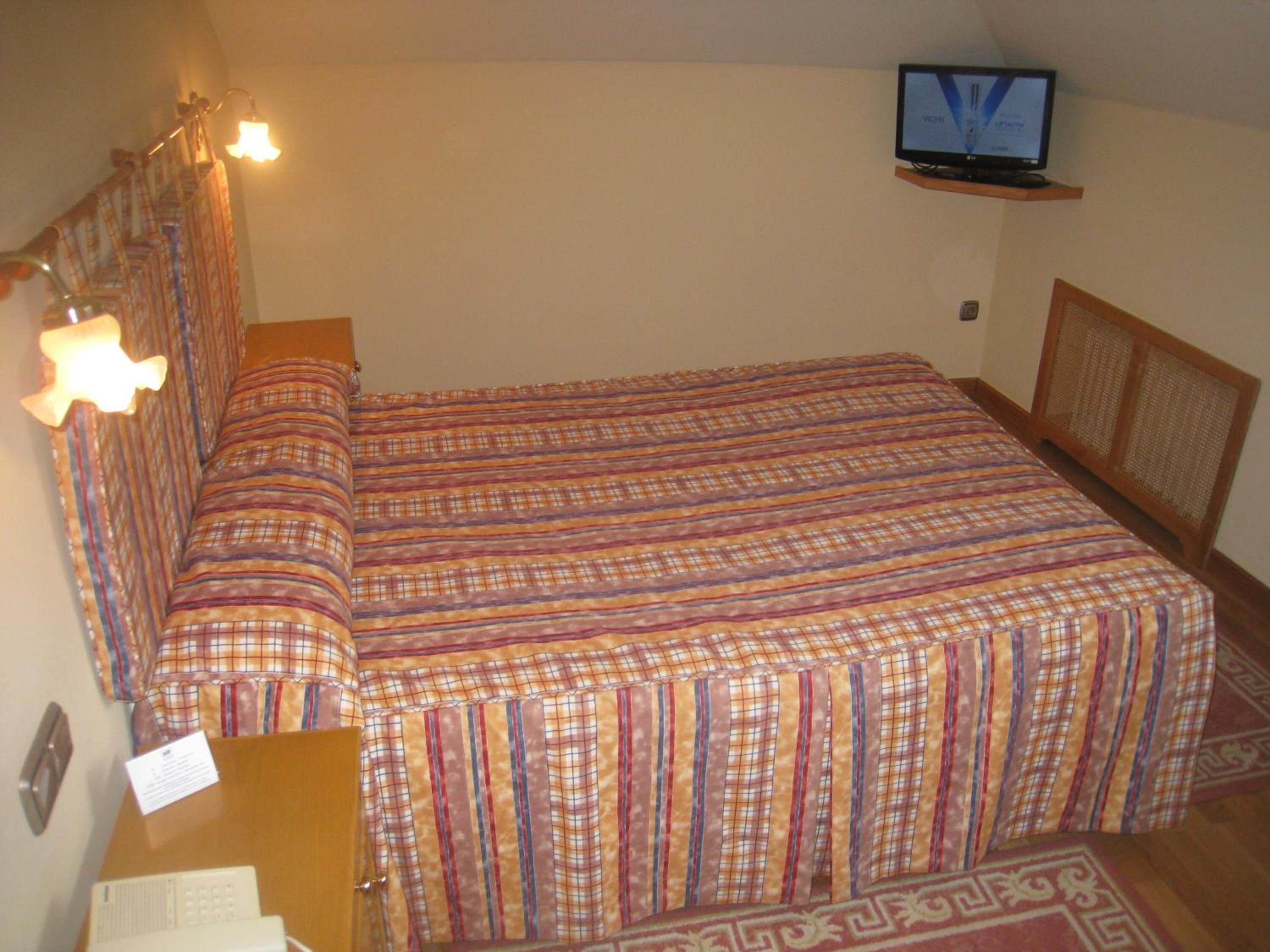 Un lit simple avec une couverture à carreaux, un téléviseur posé sur une étagère au-dessus, et un bureau avec une lampe et un bloc-notes.