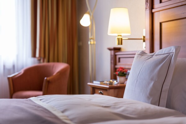 Una habitación de hotel con cama, una silla, una mesita de noche con lámpara y un ventanal con cortinas.