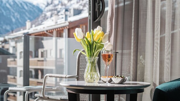Un balcón con una mesa lista para dos personas, con un florero de flores y una copa de vino, con vistas a una montaña nevada.