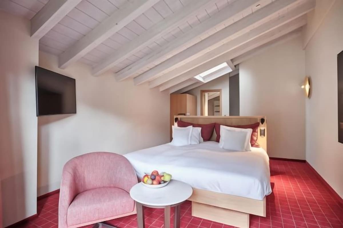 Habitación de hotel con cama, una silla, una mesita con fruta y un televisor montado en la pared.