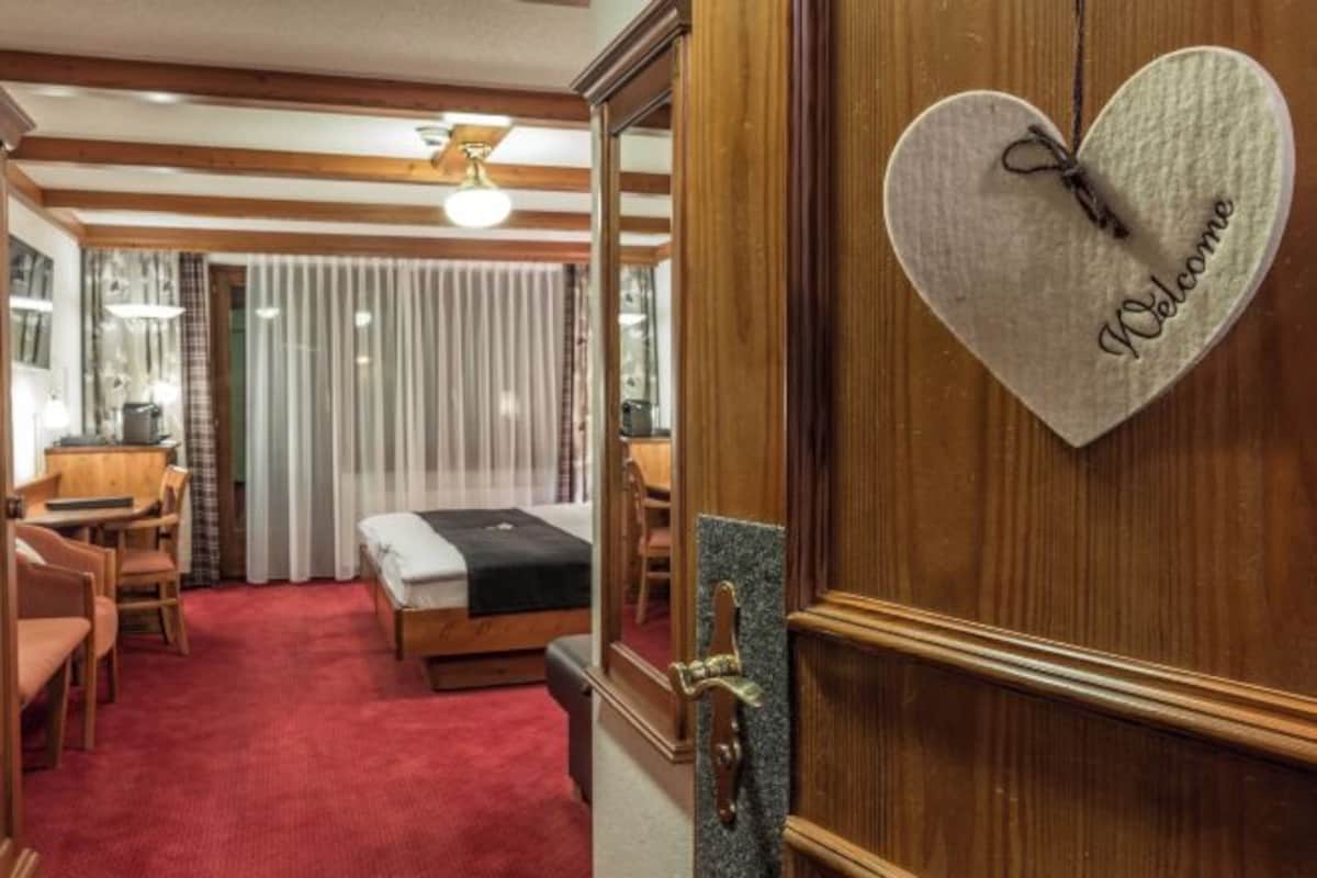 Habitación de hotel con puerta de madera, un letrero de bienvenida con forma de corazón, una cama, un escritorio y una alfombra roja.