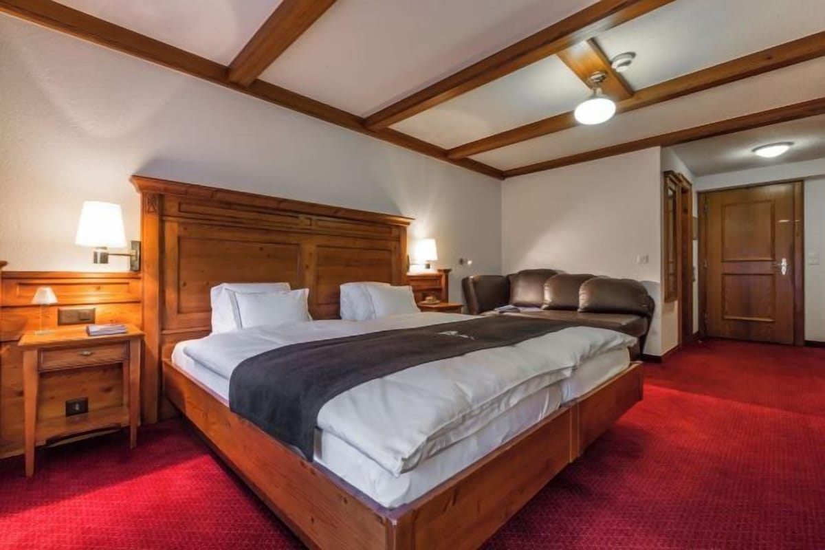 Habitación de hotel con una cama de madera, una mesita de noche con lámpara, un sillón de cuero y una alfombra roja.