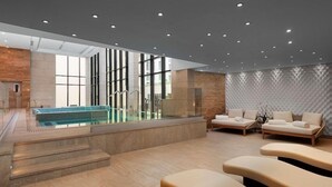 A modern indoor pool area with a clear glass wall, a few steps leading to the pool, and a seating area with cushioned loungers. - Serdika (Sofia)