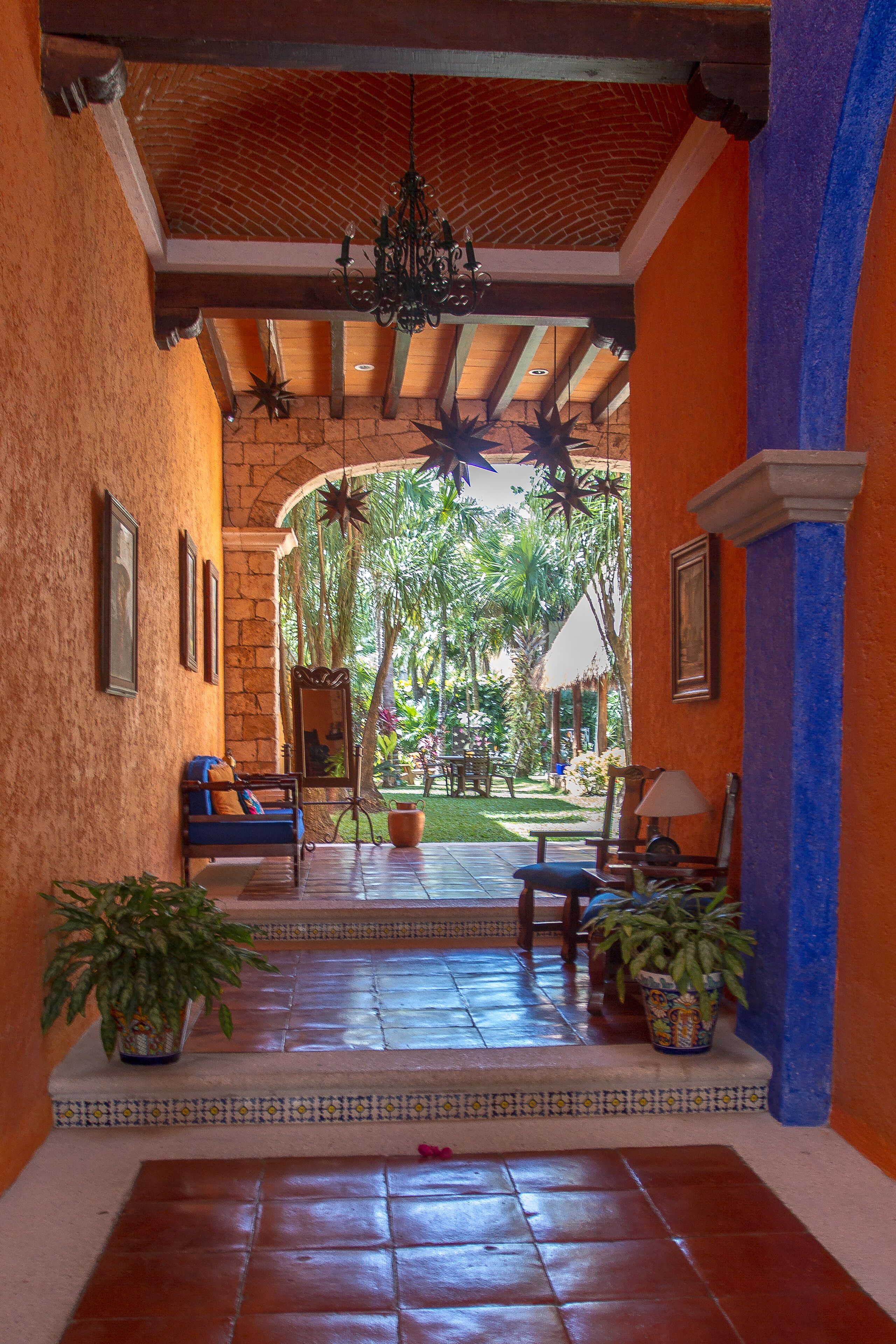 A covered outdoor area with a tiled floor, benches, potted plants, and a view of a garden.