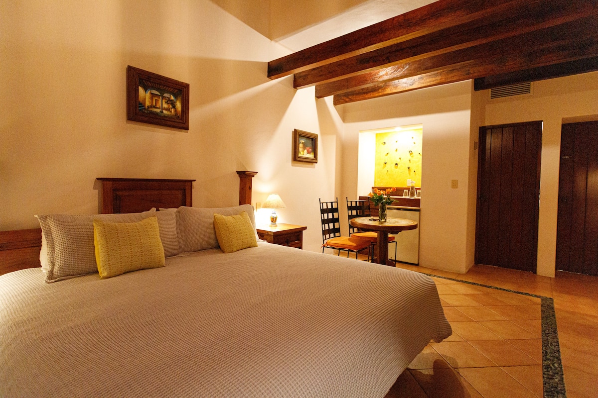 A bedroom with a large bed, a wooden headboard, a bedside table with a lamp, and a small table with chairs in the corner.