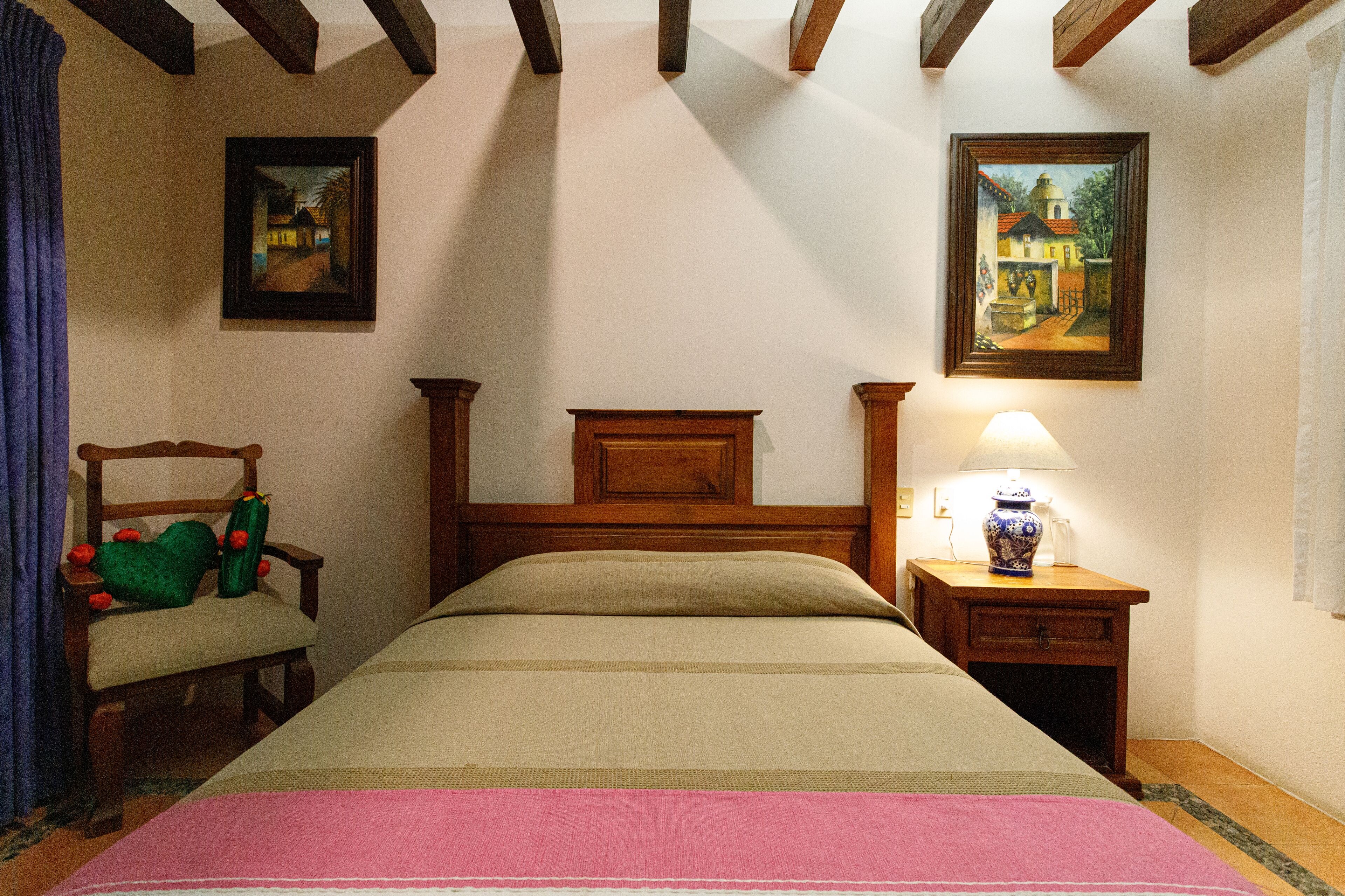 Slaapkamer met een houten bed, een nachtkastje met een lamp, een stoel met kussen, en twee ingelijste foto's aan de muur.