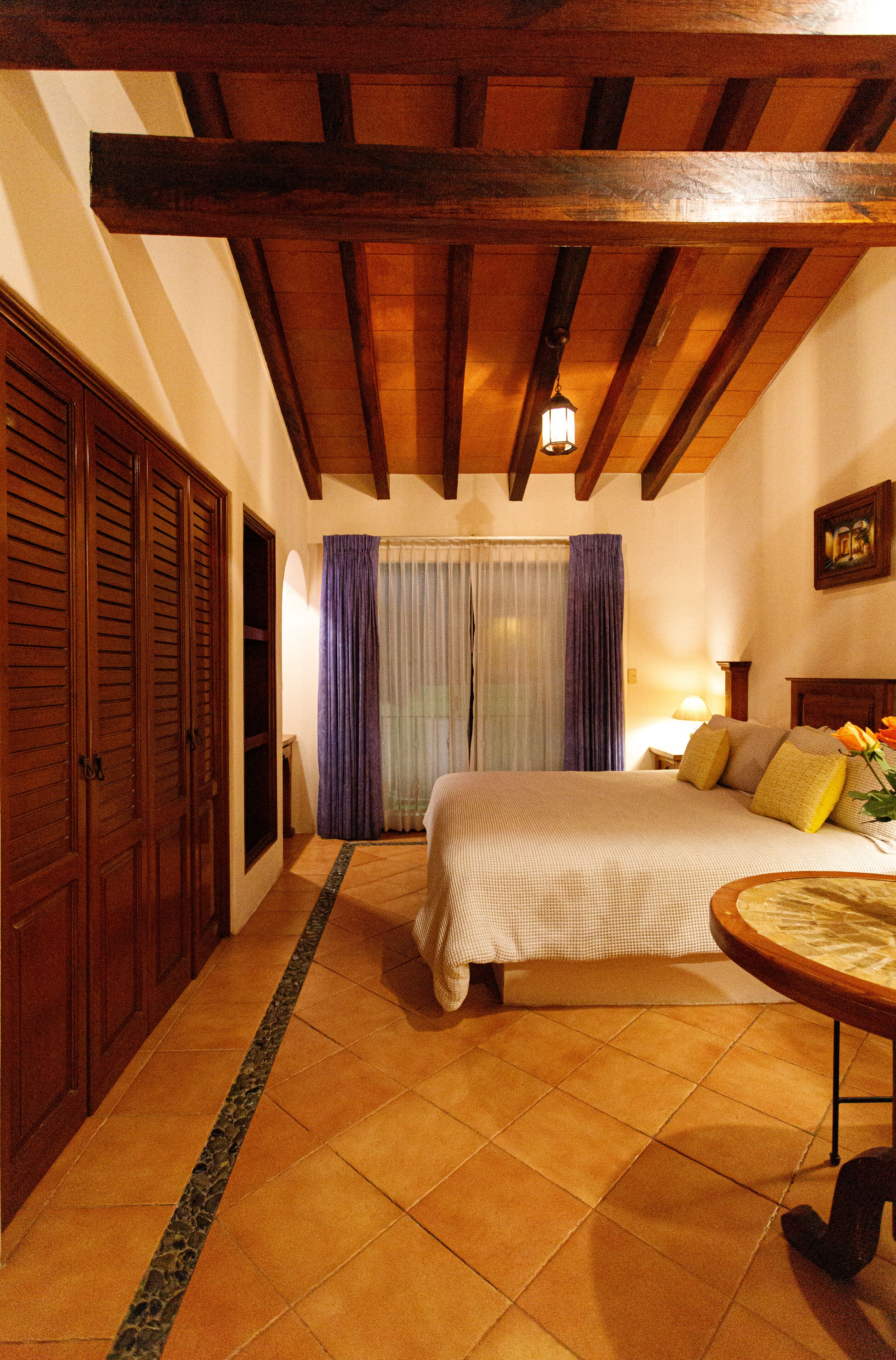 A bedroom with a wooden ceiling, a bed with pillows, a nightstand, and a window with curtains.