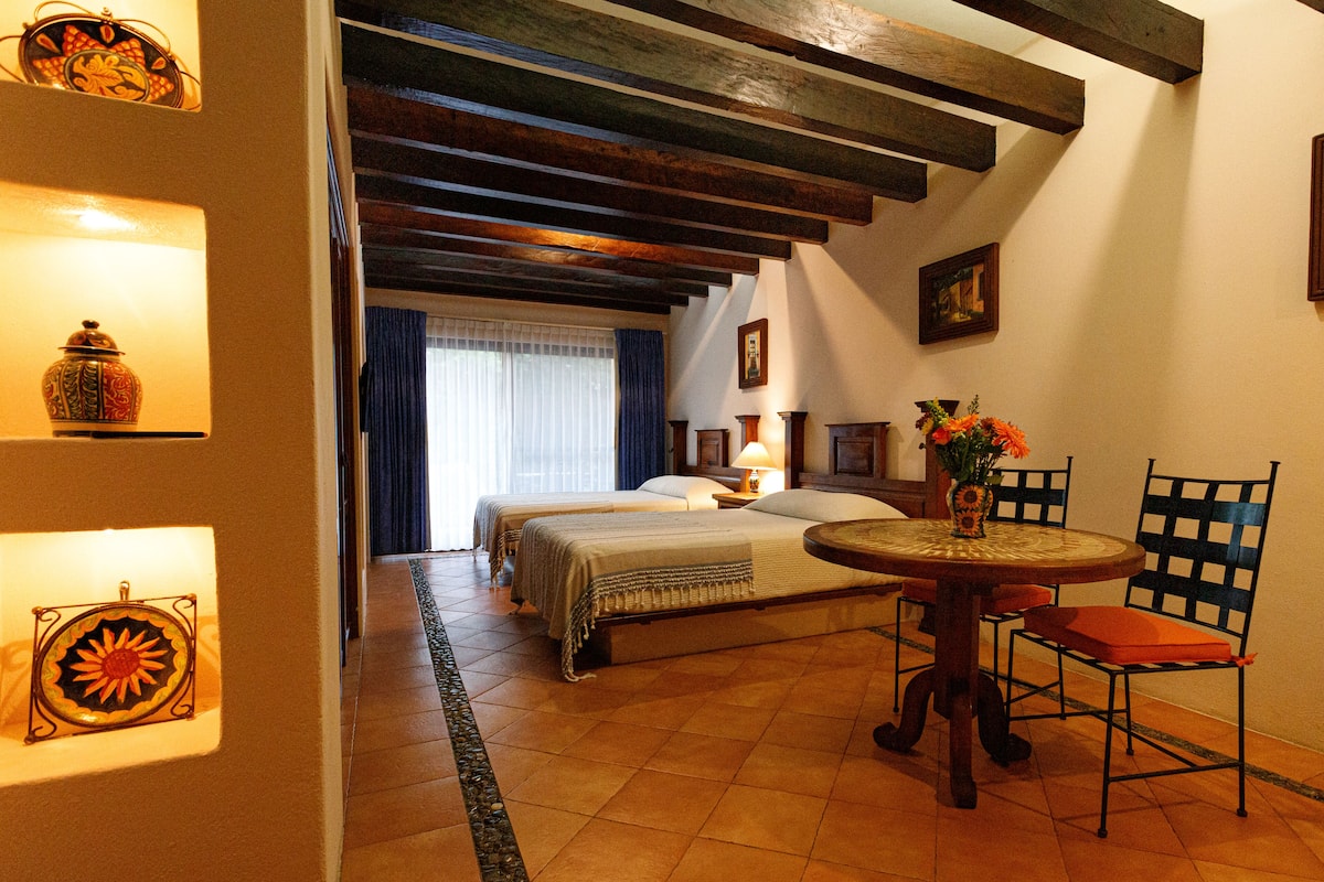 A hotel room with a bed, a dining table with chairs, a vase with flowers, and a wall-mounted picture.