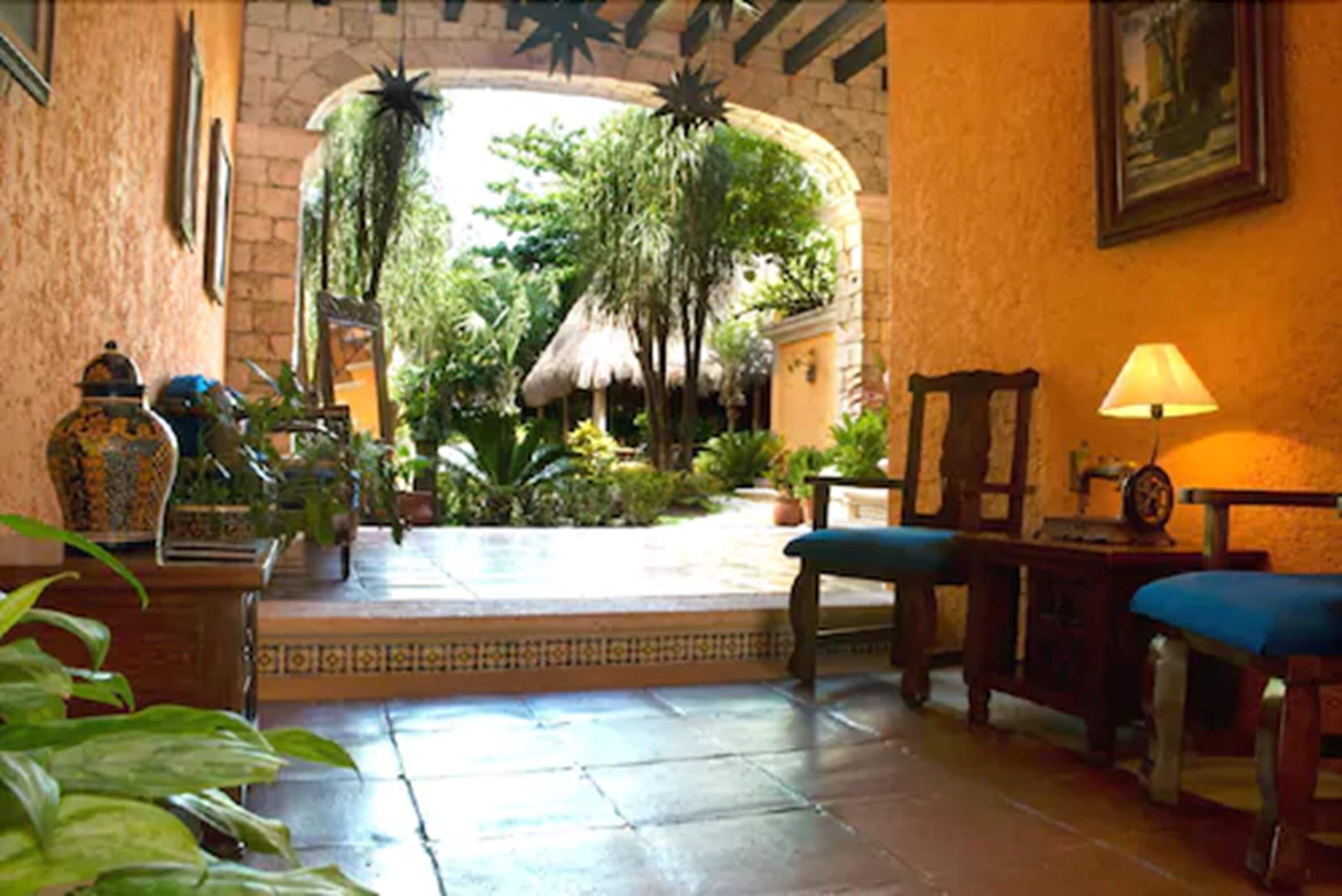 A room with a tiled floor, a wooden bench, a table with a lamp, and a view of an outdoor area with plants and a thatched roof structure.