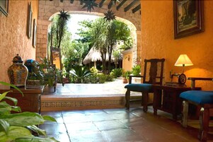 Una habitación con piso de baldosas, un banco de madera, una mesa con una lámpara y vista a un espacio exterior con plantas y una estructura de techo de paja.