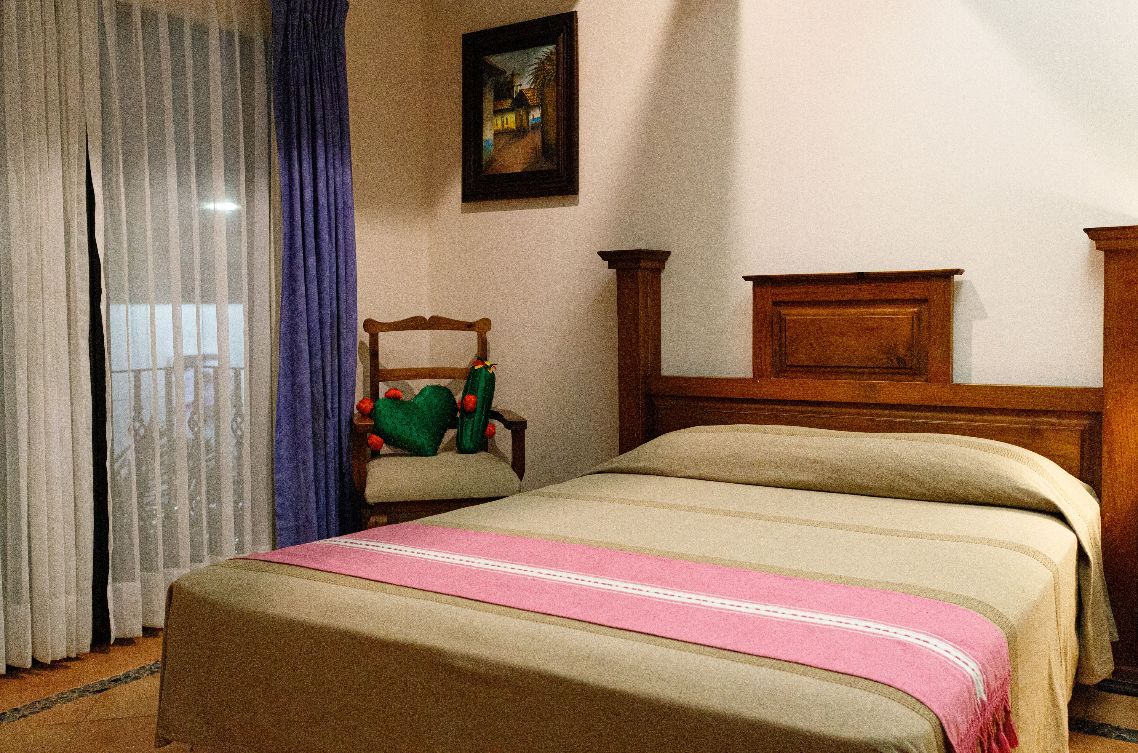 A bedroom with a bed, a chair, a painting on the wall, and a window with curtains.