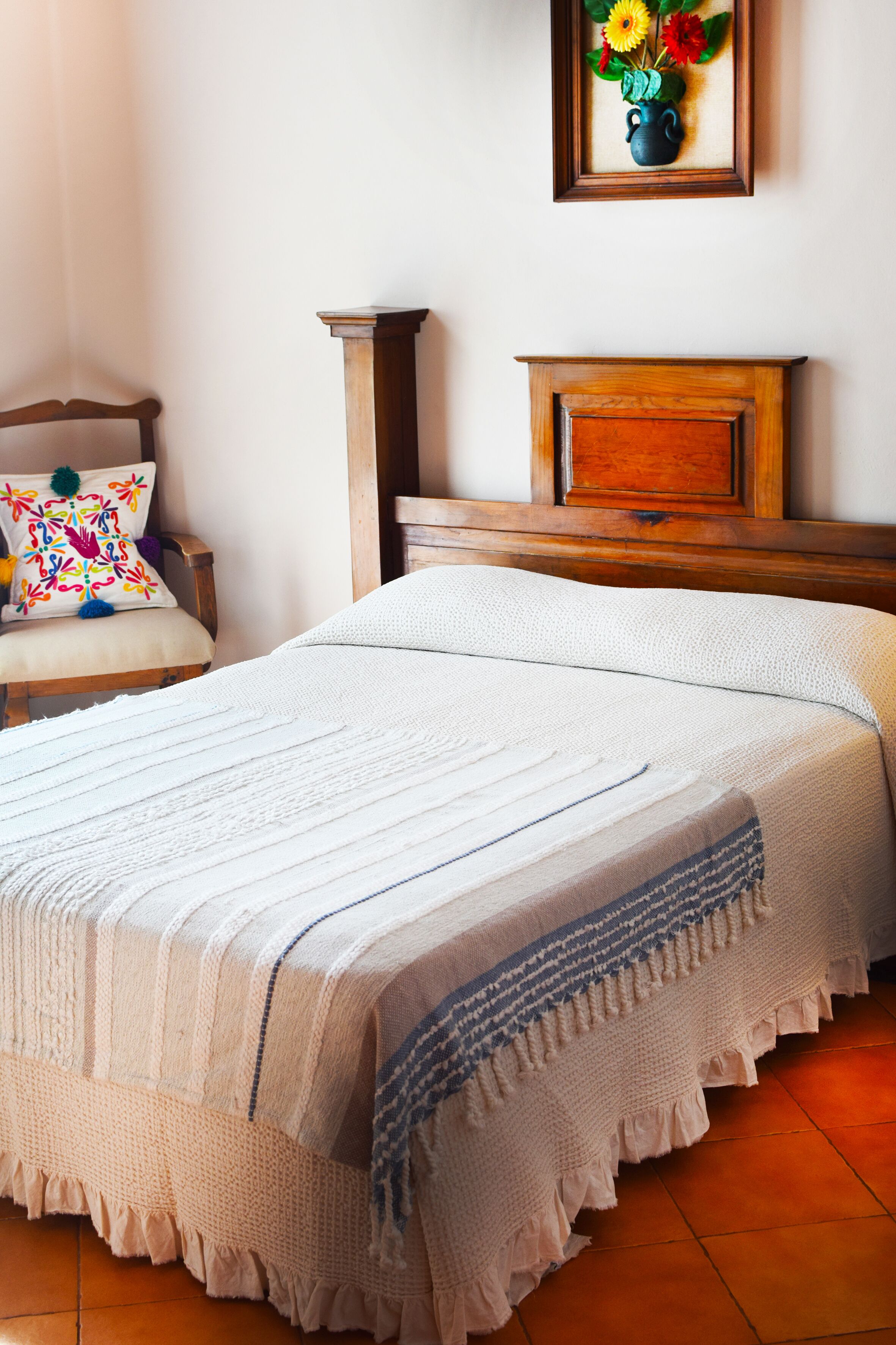 A neatly made bed with a white and blue blanket, a wooden nightstand, a chair with a colorful pillow, and a framed picture of flowers on the wall.