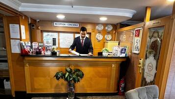 A hotel reception area with a receptionist, a reception desk, a plant, a fire extinguisher, and a chair.