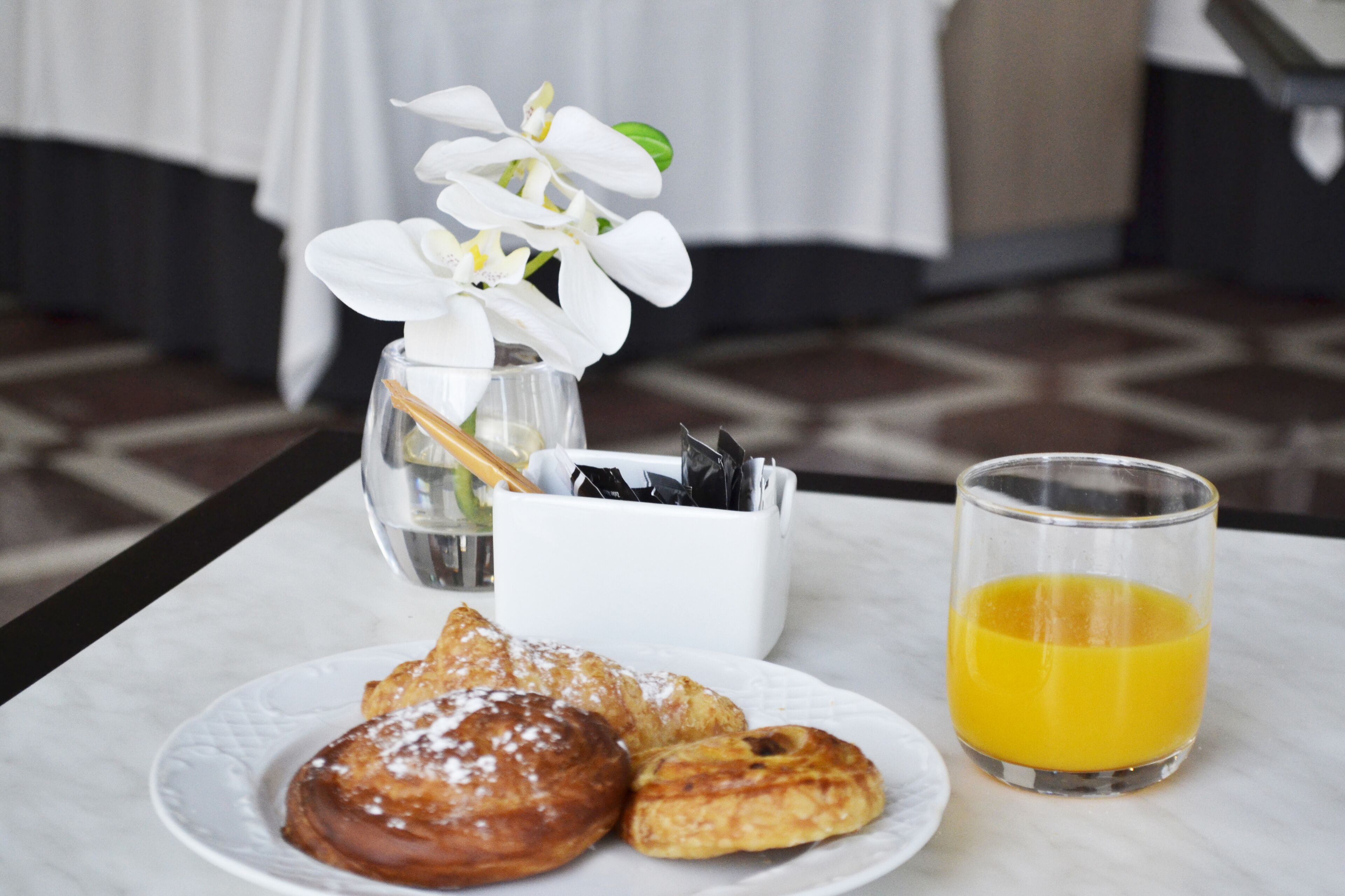 Un desayuno con pasteles, un vaso de jugo de naranja y un jarrón con flores blancas sobre una mesa de mármol.