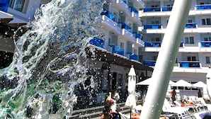 A water fountain with a green spout and a red handle, located in front of a multi-story hotel with balconies. - Hotel Acuazul (Peniscola)