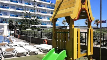 Un parque infantil con tobogán y red de escalada, con un hotel y palmeras al fondo.